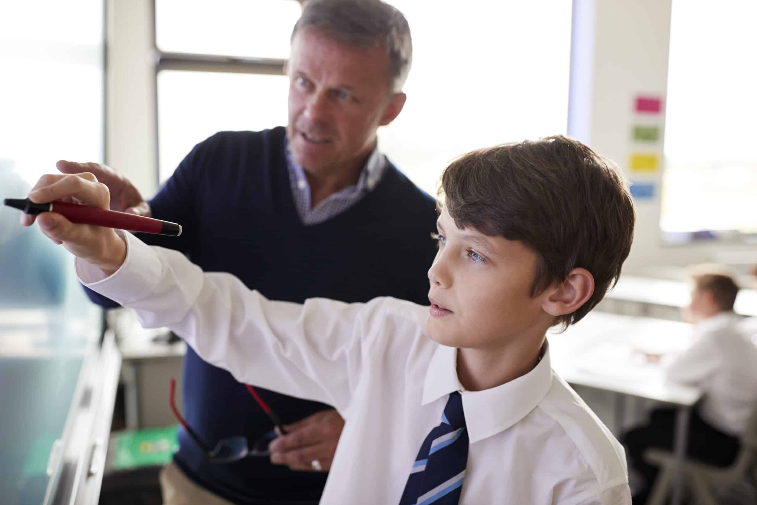 High School Teacher With Male Student Wearing Uniform Using Interactive Whiteboard During Lesson