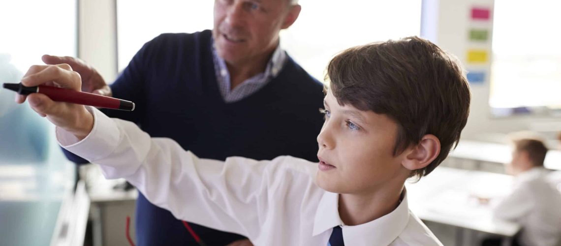 High School Teacher With Male Student Wearing Uniform Using Interactive Whiteboard During Lesson