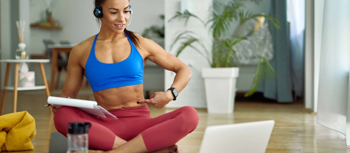 Young fitness instructor using laptop while vlogging about abs exercises at home.